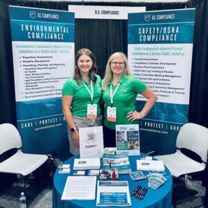 Two women in green shirts standing at a U.S. Compliance booth